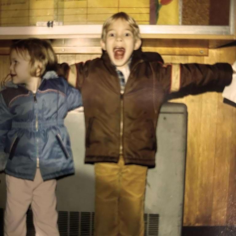 Two young children stand indoors. The boy on the right, wearing a brown jacket and tan pants, stands with arms wide open and mouth agape, excited. The girl on the left, in a blue jacket, looks away from the camera.